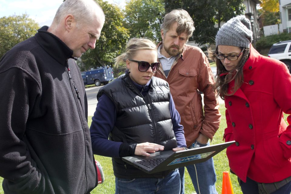 Three people stand around a woman holding a laptop while they all look at the screen with water monitoring information. 