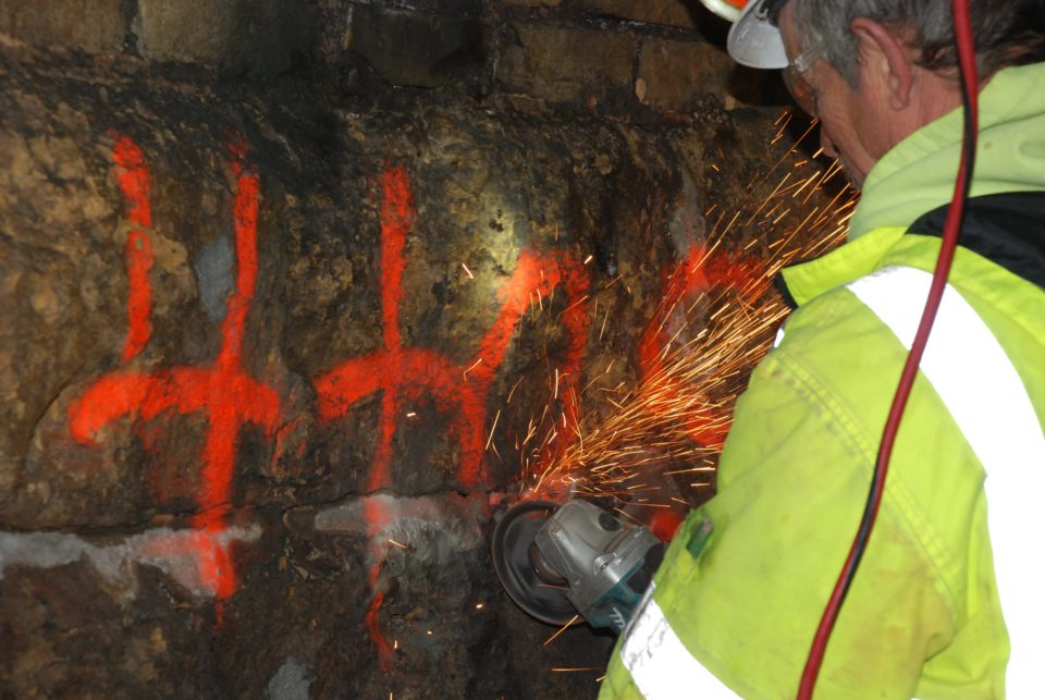 A person wearing a high-visibility safety jacket works on a part of a spray-painted stone wall with a spray of sparks flying from a handheld grinder.  