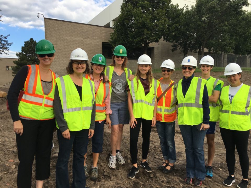 Several people smile for the camera during construction while wearing hard hats and high-visibility safety vests. 