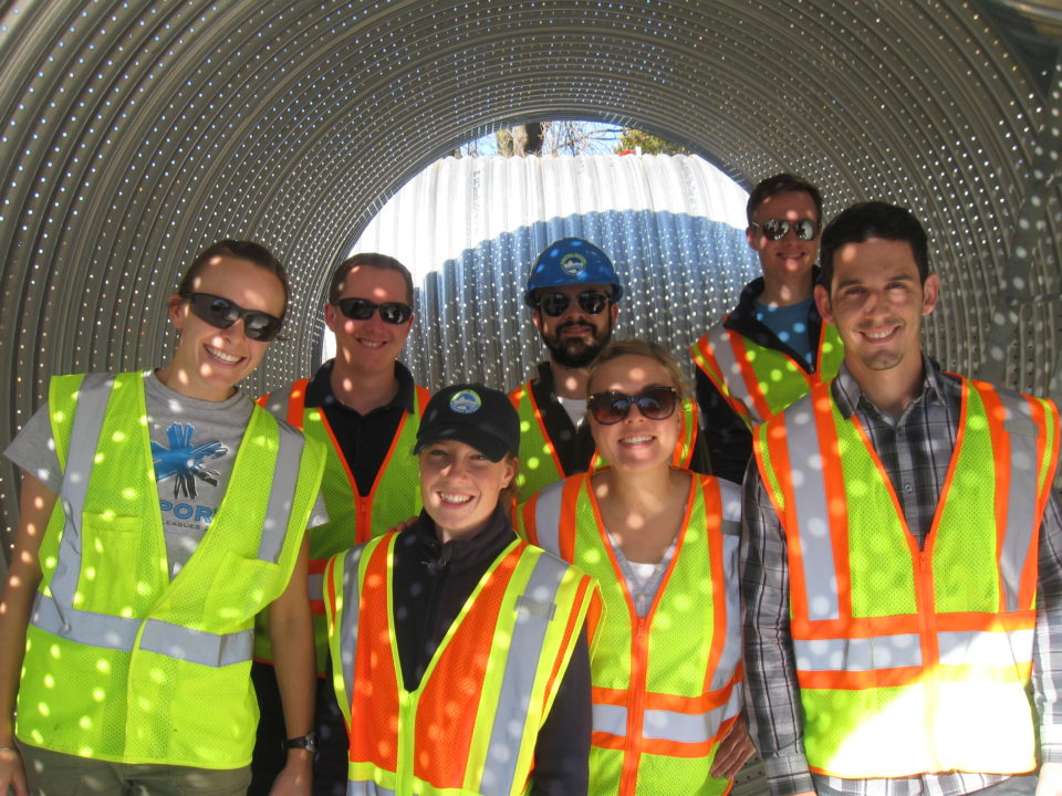 Seven smiling people wearing high-visibility safety vests stand inside a 10-foot diameter perforated metal pipe. Light shines through the holes in the pipe and shows hundreds of dots of light on the people. 