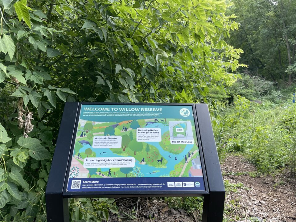 An educational sign next to a wood-chip path surrounded by green plants and trees.