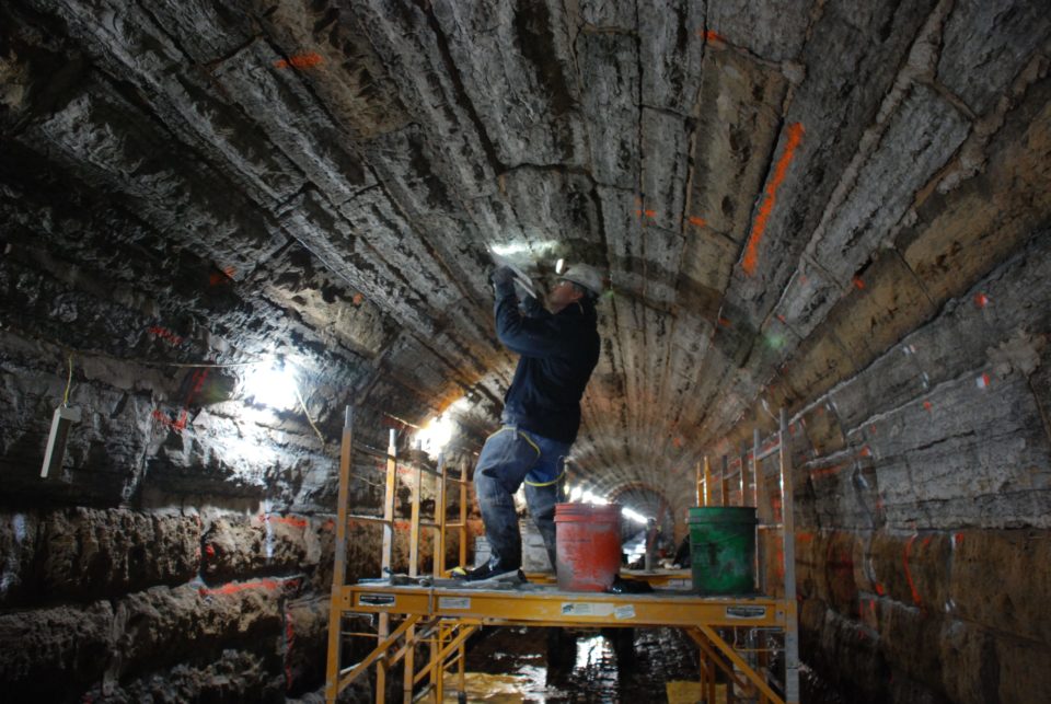 A person wearing waders, gloves and a hard hat with a headlamp works to patch the ceiling above from the top of yellow scaffolding in an arched tunnel of limestone blocks. 