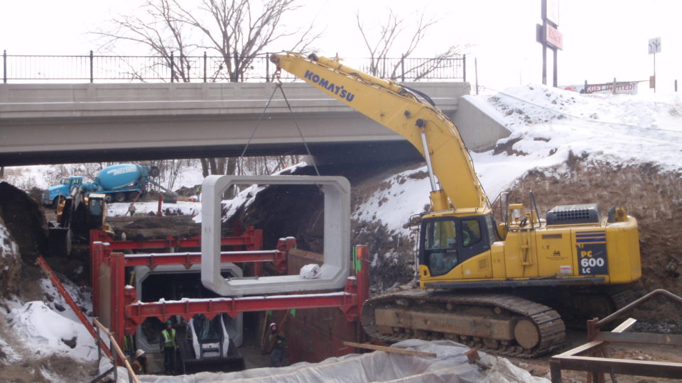 A small, yellow construction crane lifts a rectangular concrete section into place in temporary red support beams. The sections are below an overpass and snow is on the ground. 