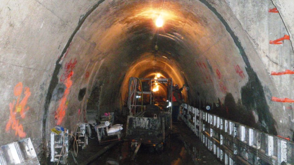 An arched underground concrete tunnel with temporary lighting. There is a scaffold in the middle and repair equipment lined up along both sides. 