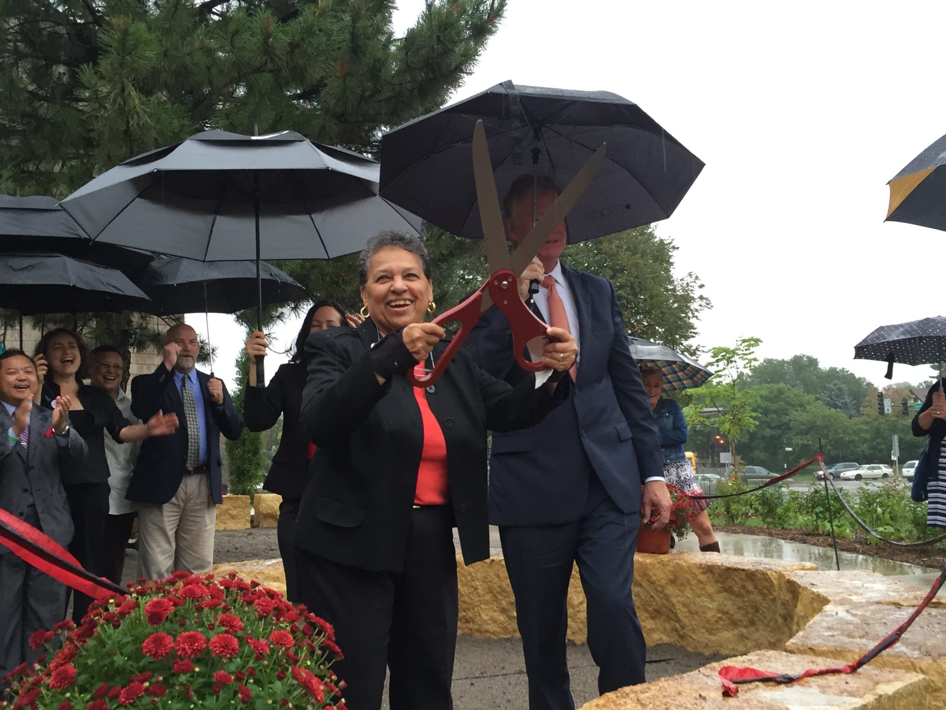 A smiling woman in business attire holds up a pair of large ribbon-cutting scissors after cutting the ribbon of a new rain garden at Central High School in Saint Paul. A crowd of people behind her are holding open umbrellas, and the stones surrounding the rain garden are wet with rain.