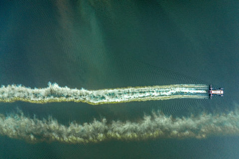 Aerial view of a boat applying alum to Como Lake, leaving a trail of white and light green stripes in the water behind the boat.