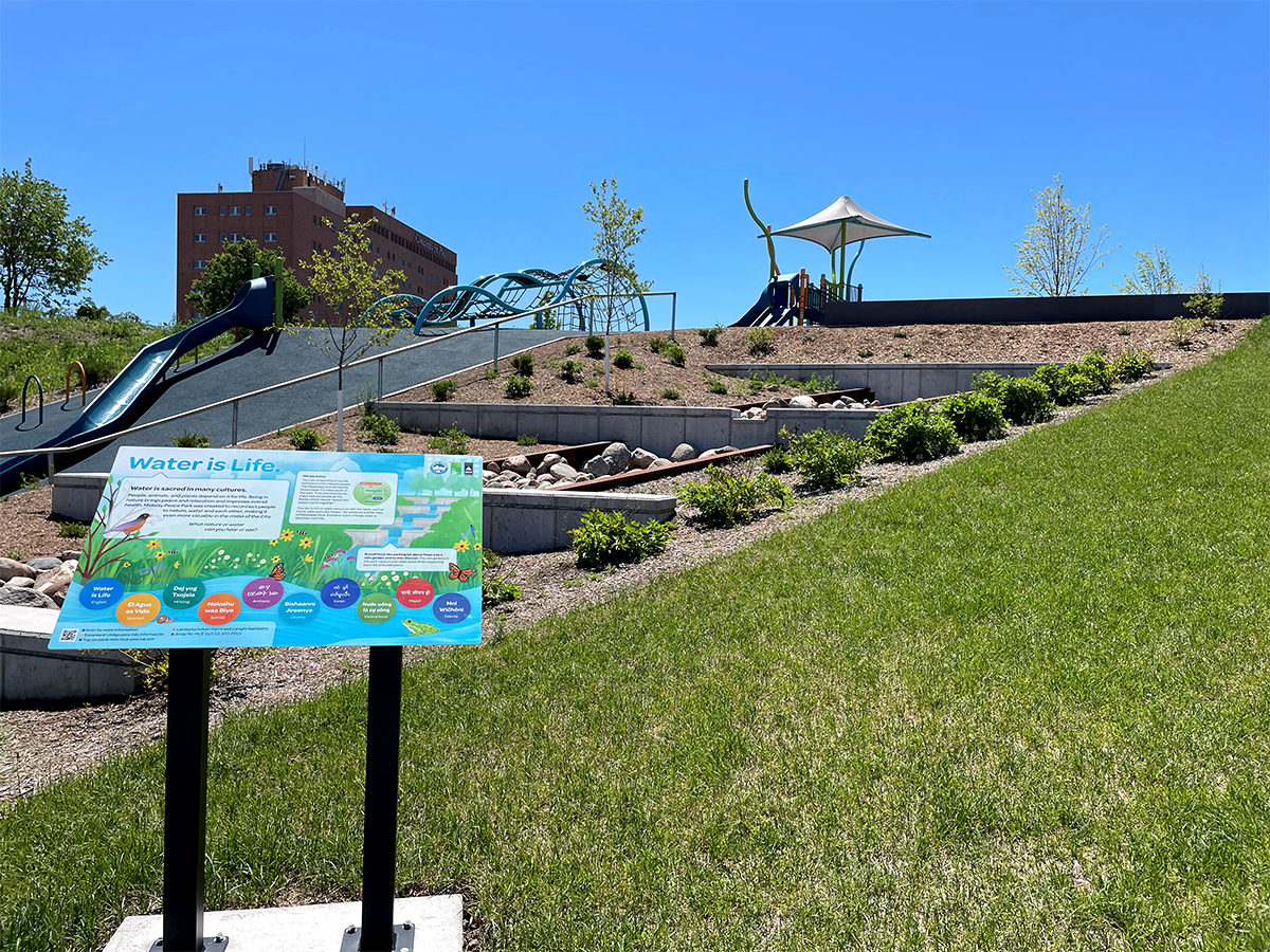An interpretive sign in front of a slope with the tiered stormwater channel and playground at the Midway Peace Park.