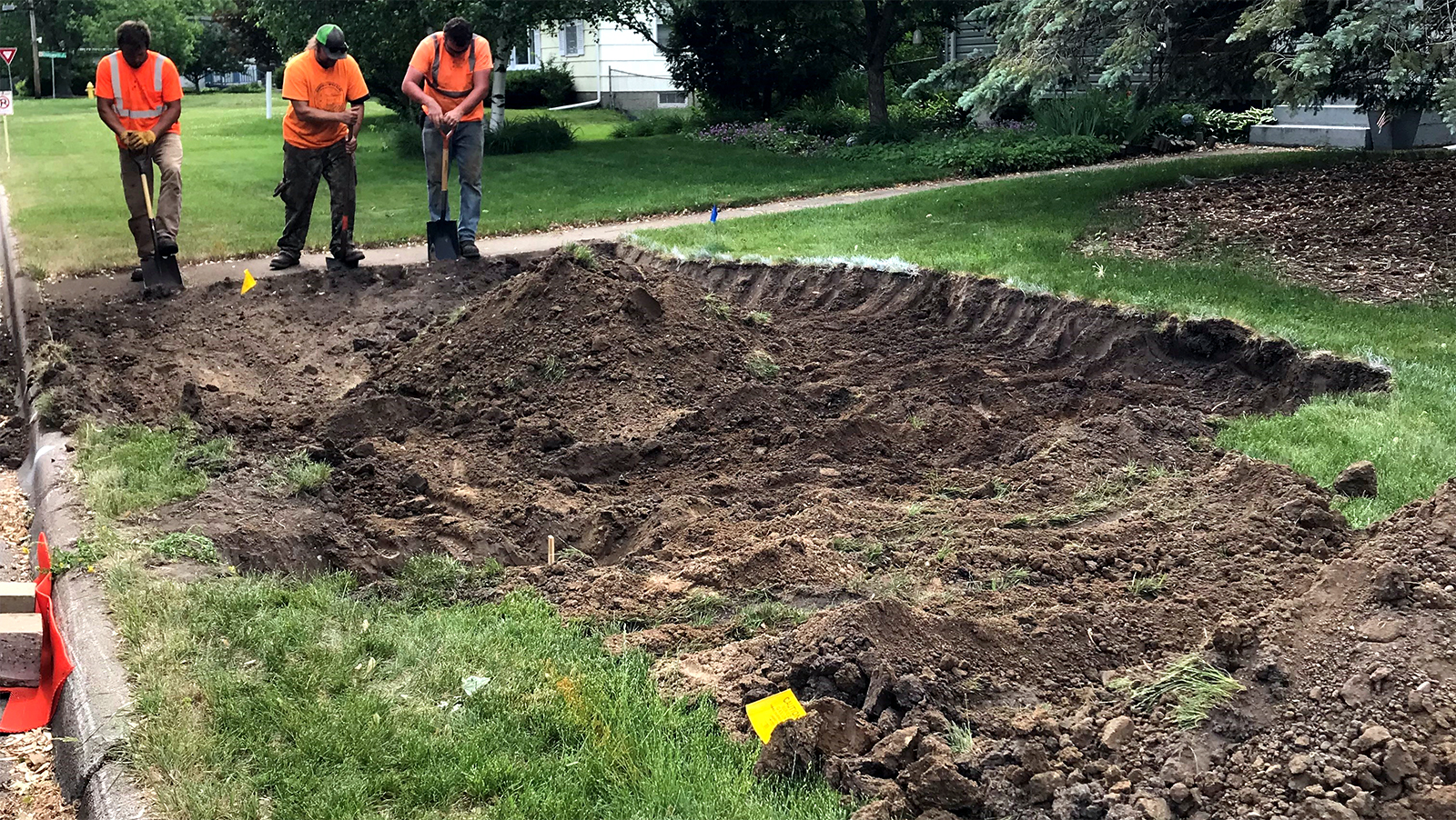 A crew wearing orange safety shirts digs a large, shallow basin in a residential lawn. Yellow flags mark underground utilities. 