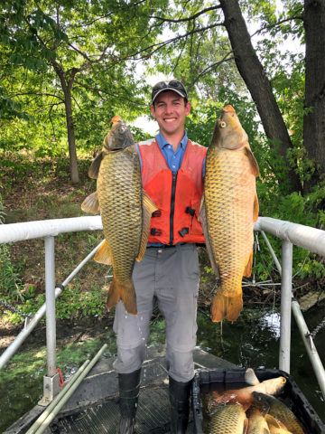 Person stands in a boat and holds up two large carp. 