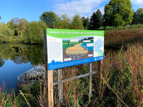 Interpretive sign next to a pond surrounded by native plants. The sign says "Ironing Out Pollution for a Cleaner Como Lake" 