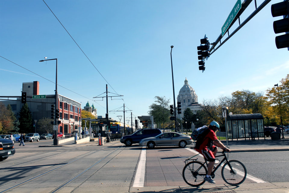 The dome of the Minnesota State Capitol building is in the background of a busy urban intersection with a person biking and cars on the street next to a bus stop, and a light rail station. 