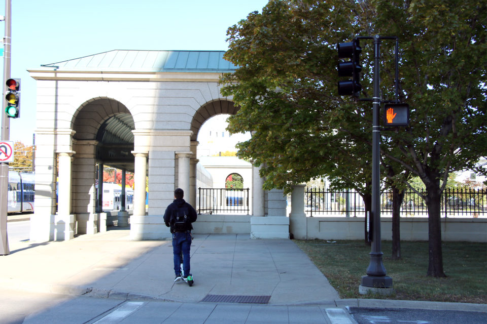 A person on an electric scooter crosses a street on the sidewalk near a METRO Green Line train. 