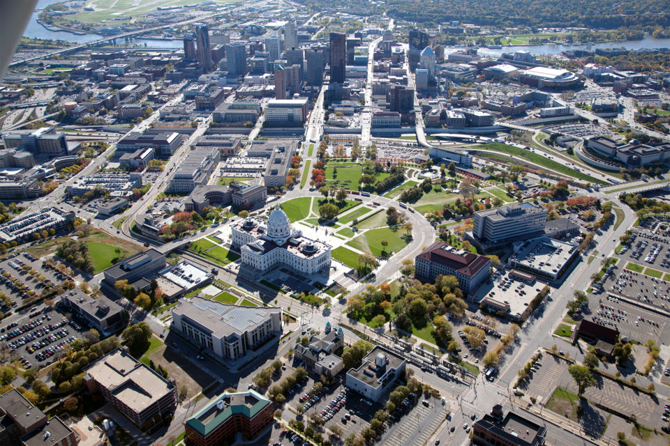 An aerial view of the Capitol Area, the Minnesota State Capitol building is in the center with the downtown Saint Paul skyline and the Mississippi River in the background. 