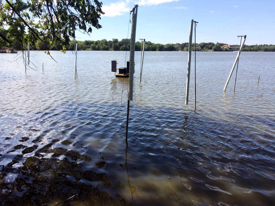 A floating box with small solar panels is next to polls connected to box nets in the lake. 