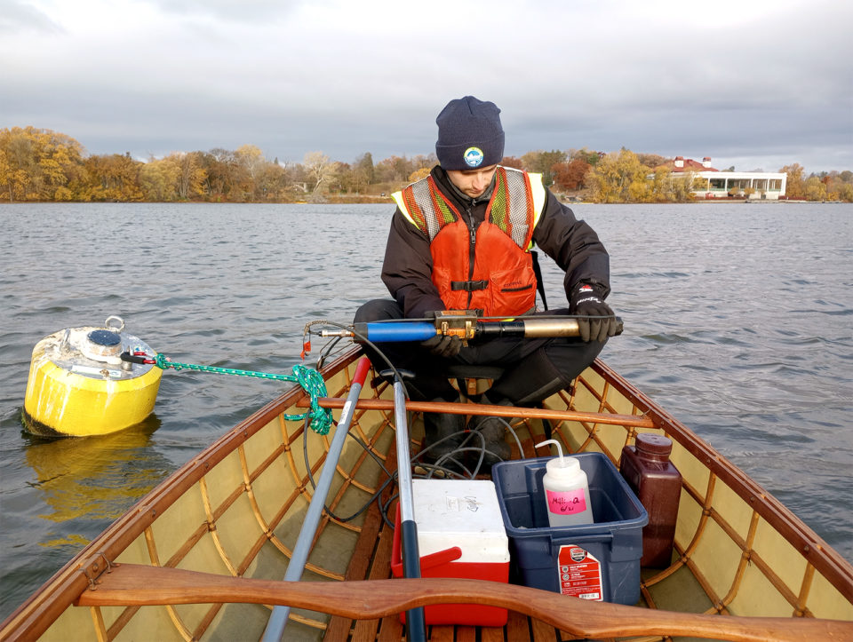 A man in a canoe holds a pipe-like instrument that is attached to a yellow buoy floating in Como Lake. 