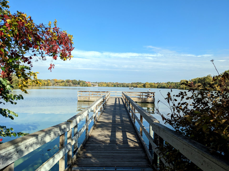 A wooden fishing pier at Como Lake, surrounded by red and maroon leaves, with a blue sky and scattered clouds. 