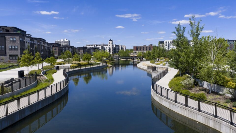 A park with a large water feature with winding banks, surrounded by walking paths and green spaces and modern apartment buildings in the background. The calm water is reflectling the blue sky dotted with small puffy clouds. 