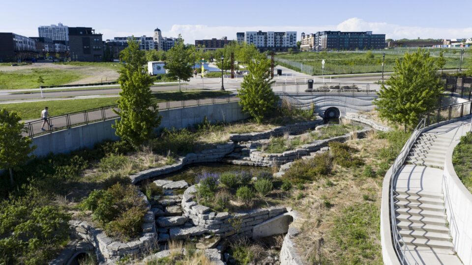 A tiered stone water feature winding through landscaped greenery in an urban park with buildings and a road in the background.