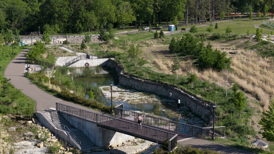 Aerial view of a scenic park featuring a winding stream channel with pathways and a footbridge crossing. People are walking along the paths and in the stream channel. 