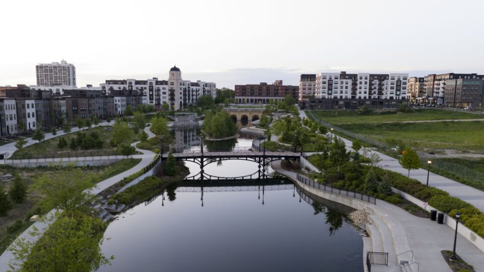 A park with a large water feature, surrounded by walking paths and green spaces and modern apartment buildings in the background. A distinctive arched bridge and pastel sunset is reflected in the calm water. 