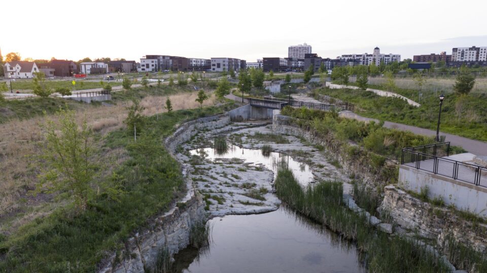 A rocky stream channel reflecting the pastel sunset, surrounded by native landscaping with pathways and modern buildings in the background. 