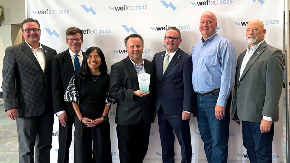 A group of seven smiling professionals proudly pose in front of the WEFTEC 2025 backdrop as the person in the center holds the Project Excellence Award.