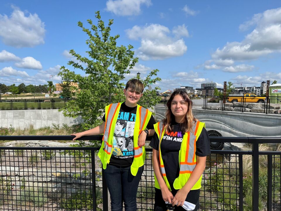 Two smiling people wearing neon orange and yellow high-visibility safety vests stand in front of a railing next to a large rain garden  