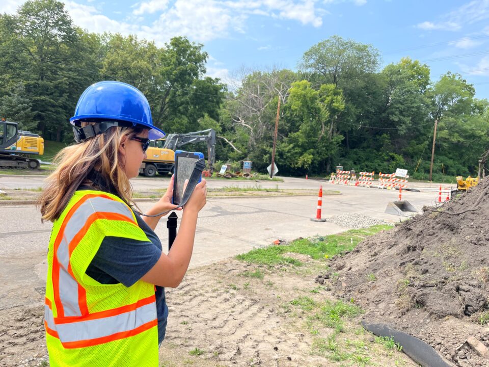 A person wearing a blue hard hat, sunglasses, and a high visibility safety vest holds a tablet to take photos. Behind her is an active construction site with orange and white cones, road work signs, and construction machinery.