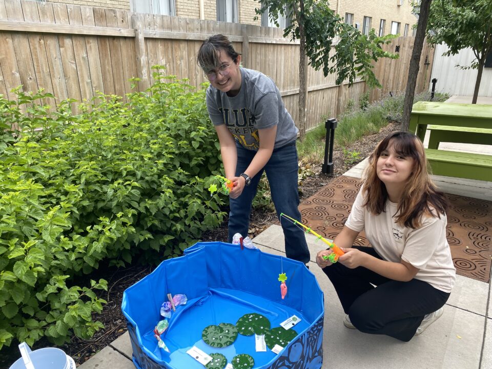 Two interns hold toy fishing poles over a small blue kiddie pool filled with fake lily pads, dragonfly larvae, and a shallow water to imitate a pond.  