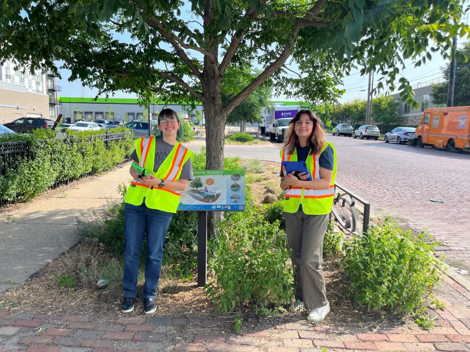 Ed and Bella wear high-visibility vests and stand in front of a rain garden and accompanying educational sign on University Avenue in Saint Paul. 