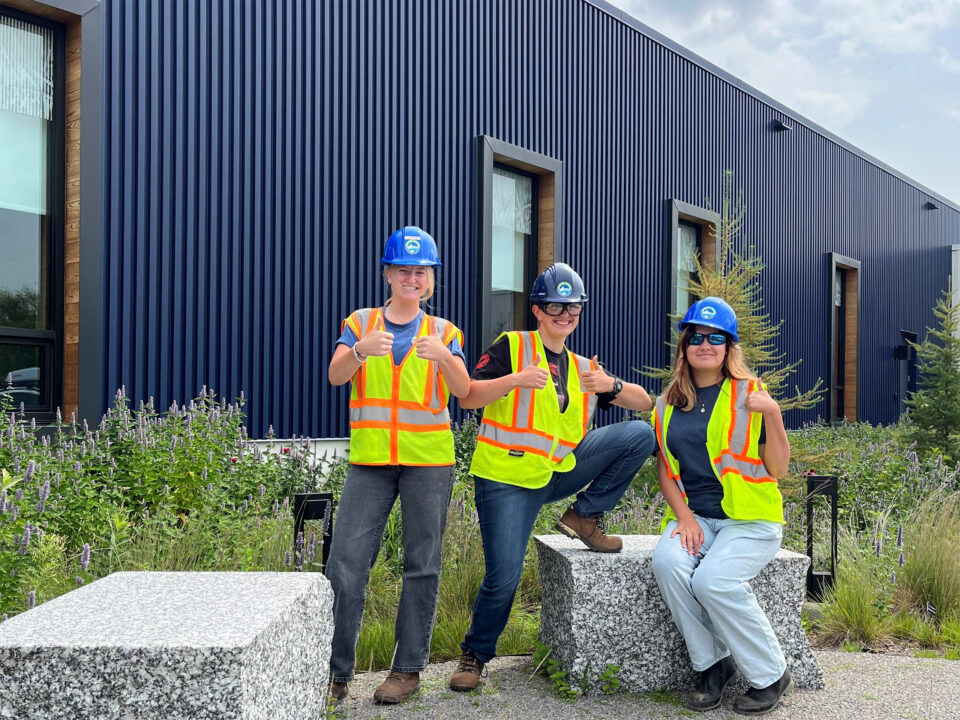 Three people wearing blue hard hats, sunglasses, and high-visibility safety vests pose for a photo before going to a construction site. They are all smiling and giving a thumbs up.