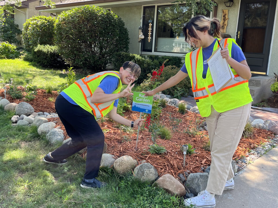 Two smiling people wearing high-visibility safety vests put a small green and blue yard sign that reads 