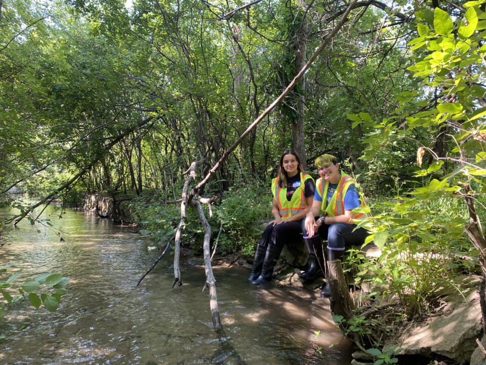 : Two smiling interns wearing high-visibility safety vests and rubber rain boots sit on a gray concrete retaining wall along a creek with their boots in the flowing water. Trees and vegetation surround them.
