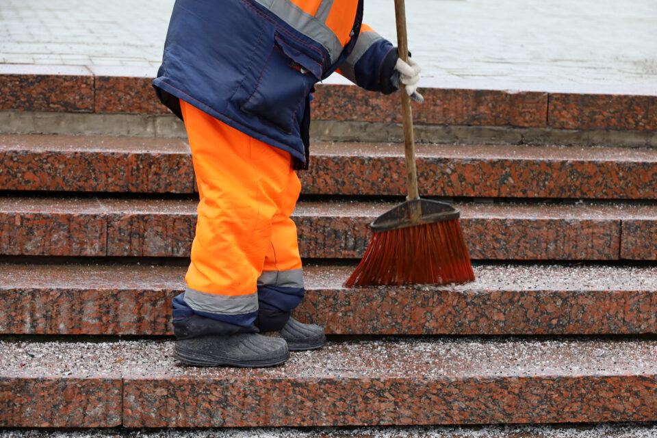 A worker wearing a winter maintenance uniform with bright orange snow pants uses a red broom to sweep granite stairs. 