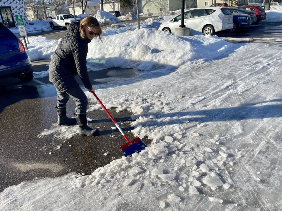 A person wearing sunglasses and a winter coat uses a red, long-handled, heavy-duty snow and ice scraper to break up compacted snow in a parking lot. They cast a long shadow in the bright afternoon sunshine.