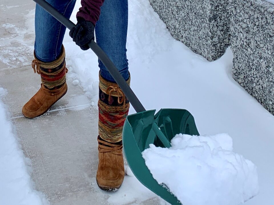 A person wearing knee-high snow boots uses a shovel to remove snow from a sidewalk.