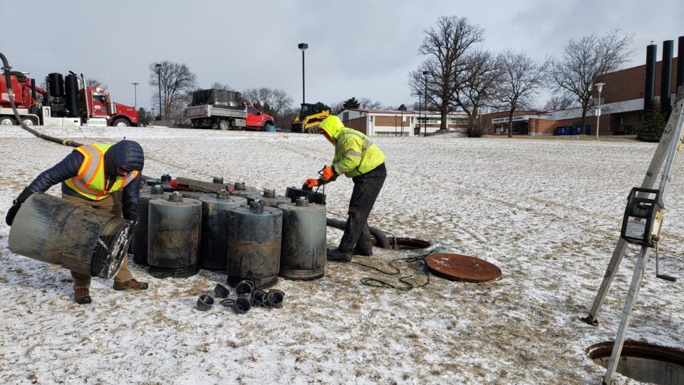Two people work on several large, black cylinders on a lightly snow covered hill with a school building in the background.  