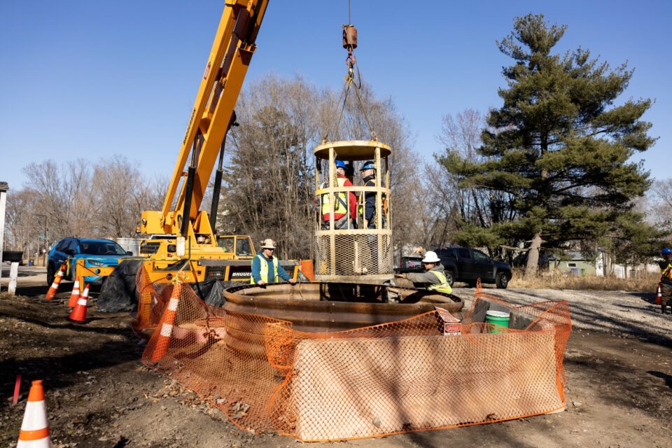 Four people in safety gear stand in cylindrical basket being lowered by a crane into a large corrugated metal tube sticking up from a street.