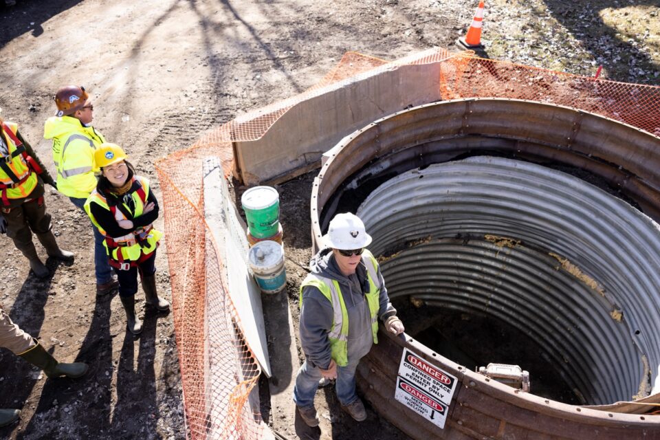 A person in a hard hat and safety vest stands next to large rings of corrugated metal that lead to a storm sewer tunnel under the street. The rings have warning signs on them and are surrounded by orange construction fencing.