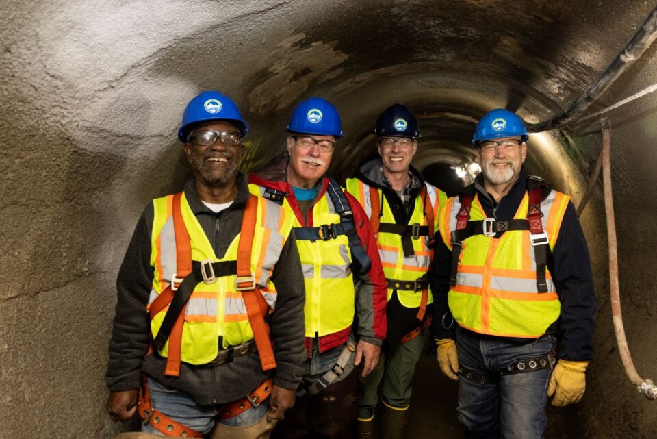 CRWD Board Managers Rick Sanders, Joe Collins and Shawn Mazanec stand in an 8-foot diameter concrete storm sewer with Administrator Mark Doneux. They are wearing hard hats, eye protection, and safety vests and harnesses. 