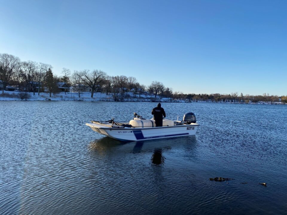 A person standing in a motorized boat with two large containers in it on a lake. There is snow covering the far lakeshore and the trees don’t have leaves on them. 