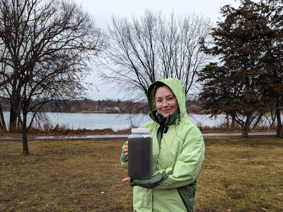 A woman wearing a light green raincoat holds a transparent jug filled with dark brown stormwater. She is standing in an area with trees and grass with Como Lake behind her. 