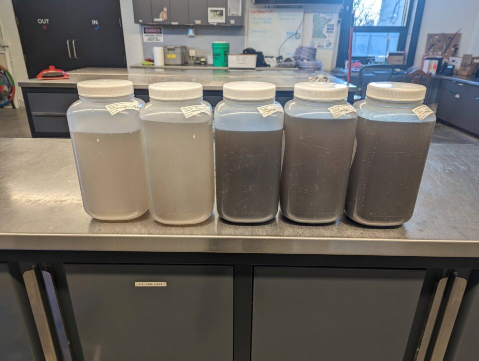 Five transparent jugs on a metal-topped table in a work area. The three jugs on the right are filled with dark brown stormwater, and the two on the left have light brown stormwater. 