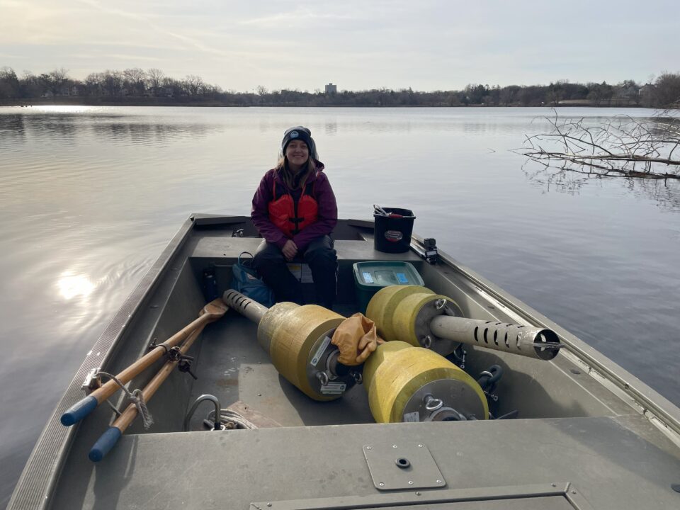 A woman wearing warm clothes and a life jacket sits in a flat-bottomed, metal fishing boat on a calm lake in early spring. Three large yellow buoys lay on the bottom of the boat in front of plastic bins with equipment. 