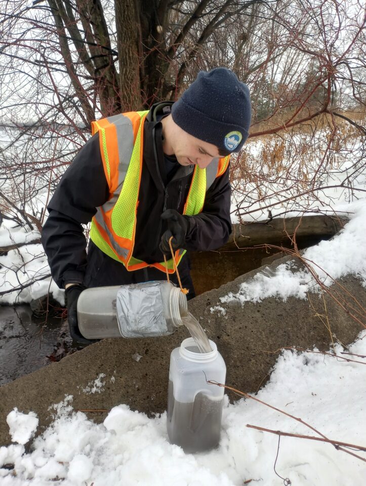 man wearing a high-visibility safety vest pours dirty, dark brown water from one transparent jug into another one sitting in snow. The man is standing along a low concrete wall of a storm sewer outfall to Como Lake. Snow on the ground, and trees are in the background.