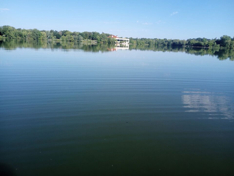 Como Lake with peaceful glassy water with small ripples. The Como Pavilion and trees on the shore are reflected on the far side of the lake. 