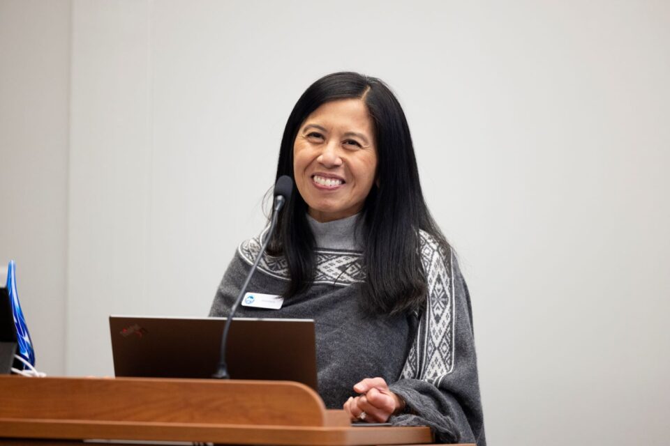 Anna Eleria stands smiling at a podium with a microphone and a laptop, in a room with a plain background. She is wearing a gray sweater with a geometric pattern.