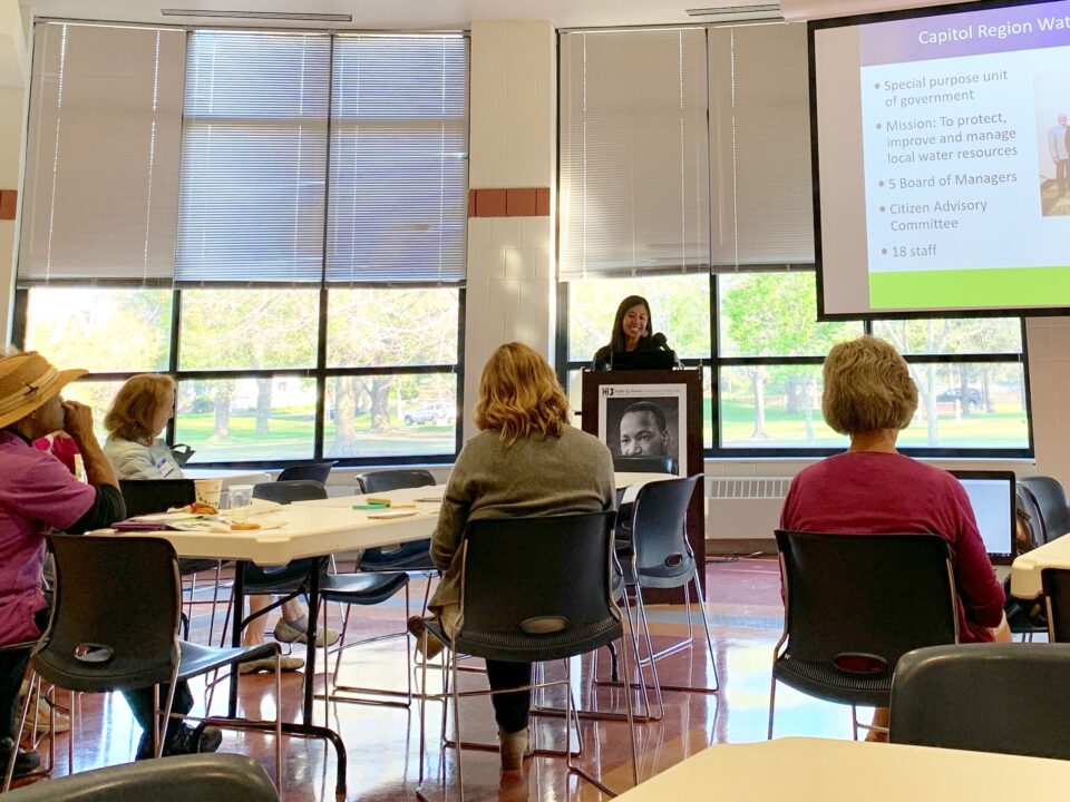 Anna Eleria is speaking at a podium with audience members seated and listening, and a slide detailing organizational information is displayed in the background.
