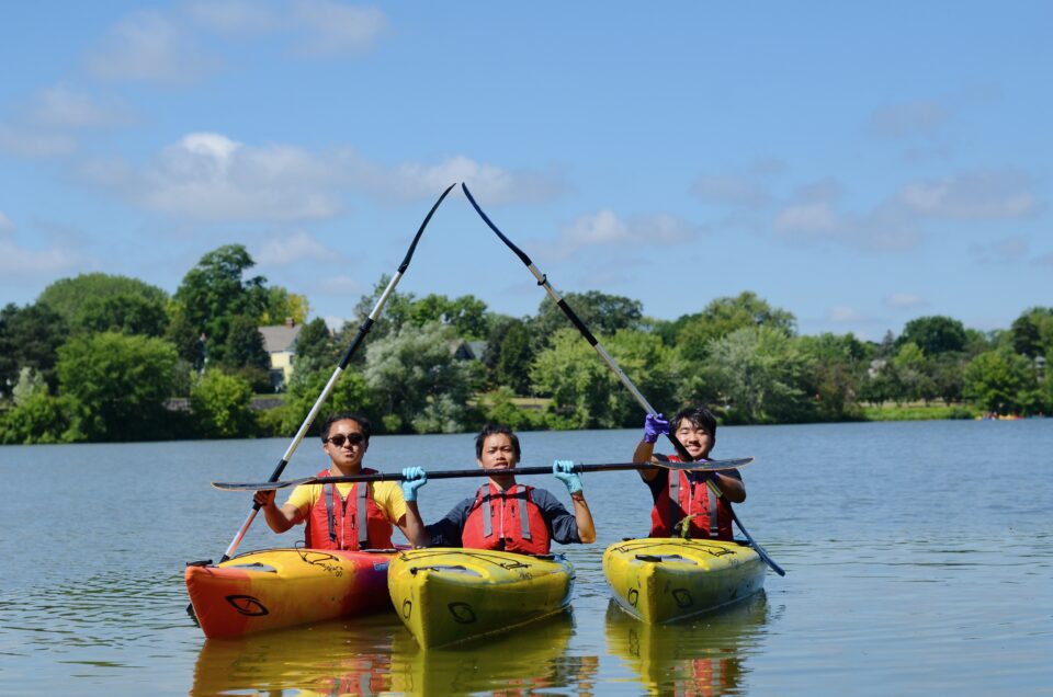 Three people wearing red life jackets on a calm lake on a sunny day, each in a separate yellow kayak, holding paddles above their heads in a celebratory gesture.