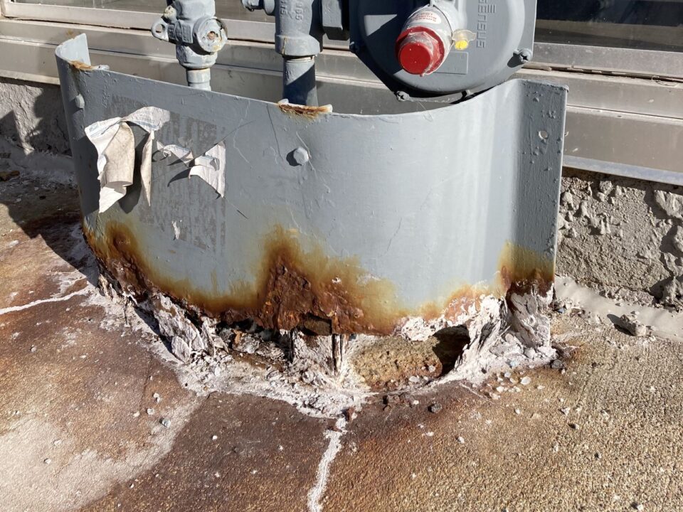 Rust and corrosion damage on a gray metal utility box and concrete sidewalk. The sidewalk is scattered with salt granules. A gas meter and piping are attached to the box.